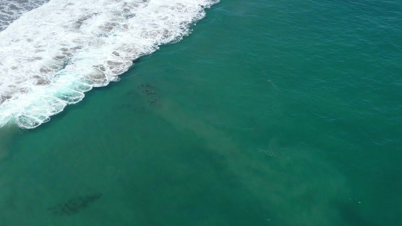 Aerial frame over San Jose del Cabo water where white wave foam spreads farther along the upper left above green-blue ocean, with no visible people.