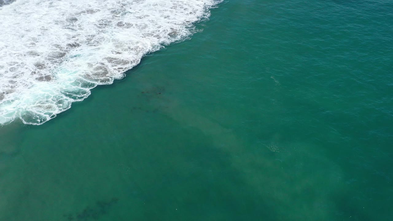 Top-down aerial shot near San Jose del Cabo with foamy surf occupying more of the upper left corner and teal ocean filling the lower frame, with no visible people.