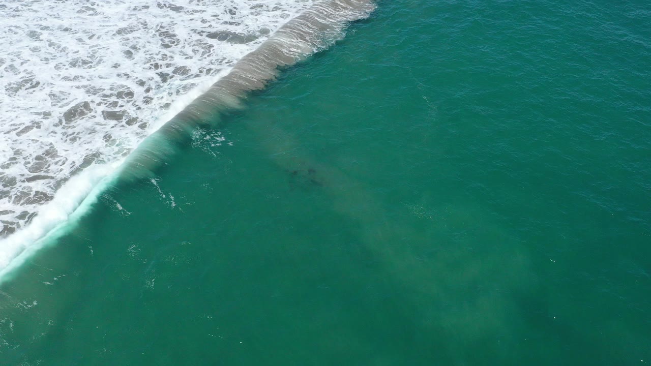 Aerial view over green sea water in San Jose del Cabo, Mexico, with foamy white surf cutting diagonally across the upper left and no visible people.