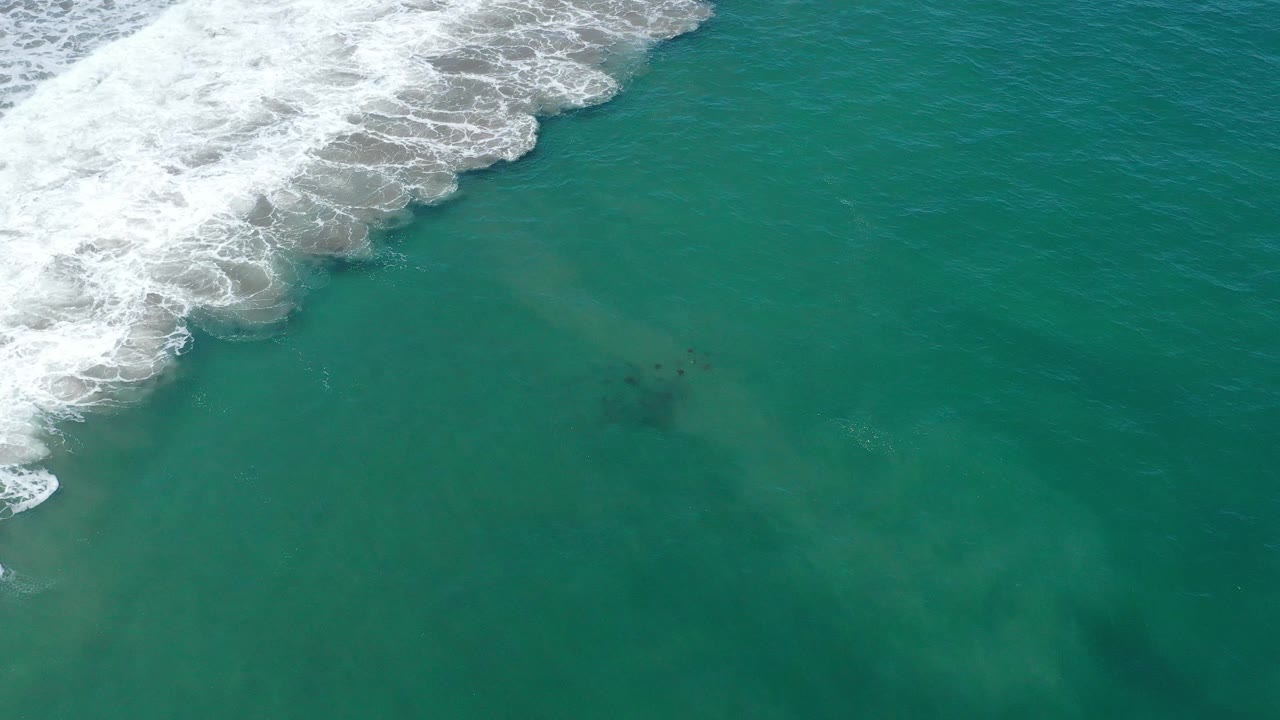 Top-down coastal water in San Jose del Cabo, Mexico, with white surf hugging the upper left corner and a faint sandy cloud below the surface, with no visible people.
