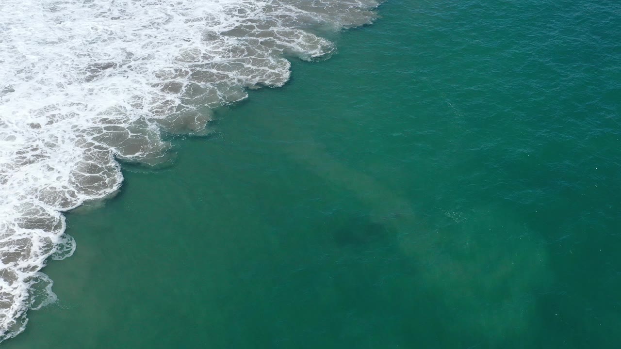 High overhead view of San Jose del Cabo ocean with foamy surf receding toward the upper left and darker teal water across the lower frame, no visible people.