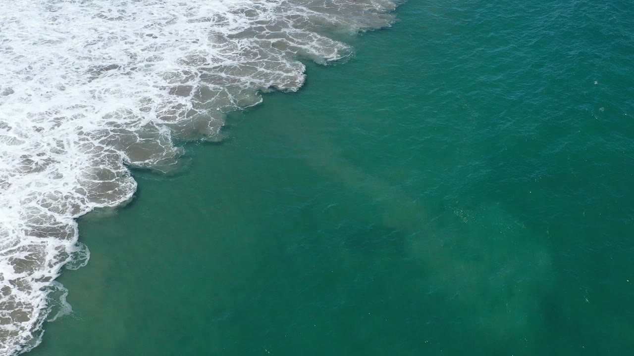 Overhead San Jose del Cabo sea frame with a diagonal strip of white surf along the left and upper areas, teal water below, and no visible people.