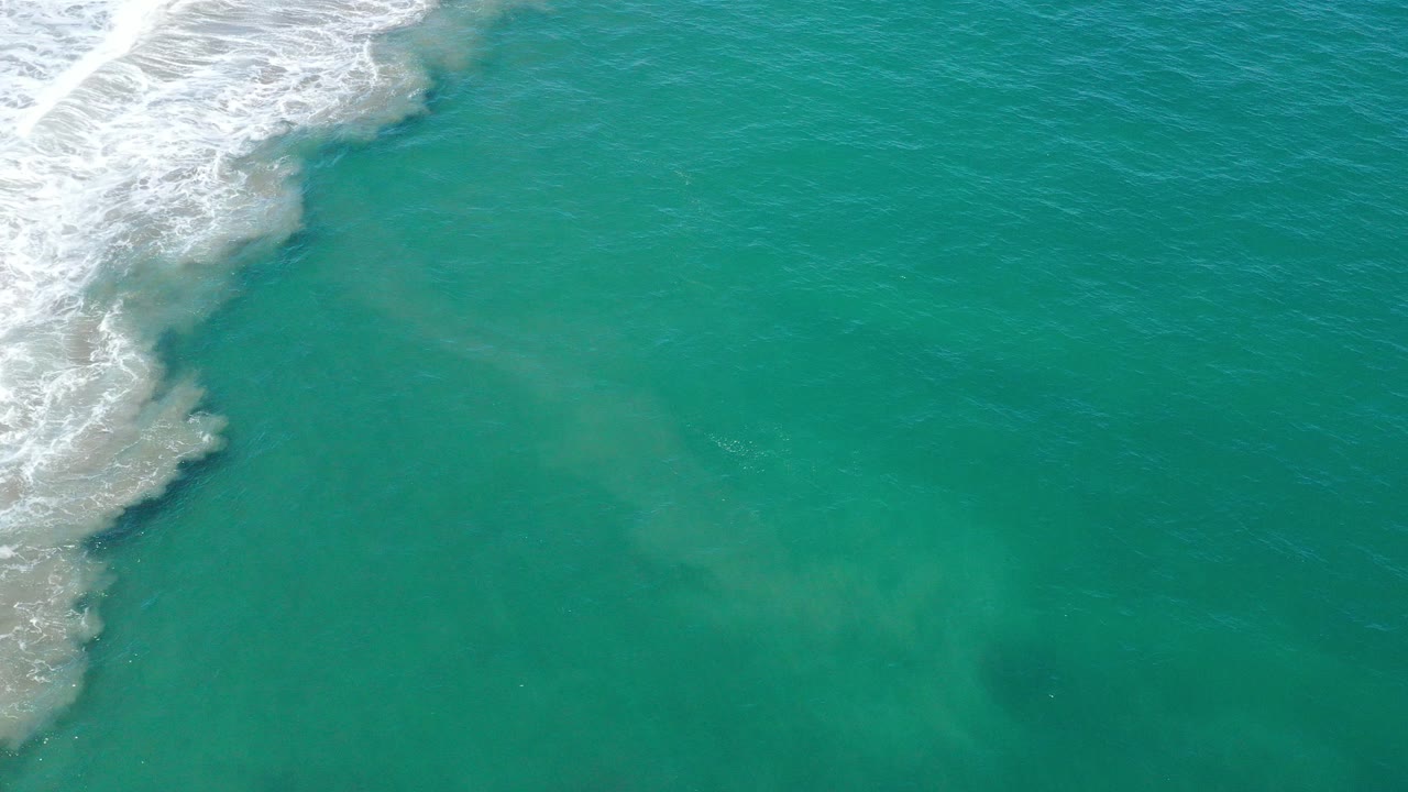 High overhead view of blue-green ocean in San Jose Del Cabo, Mexico, with white surf foam pressing in from the upper left and no visible people.