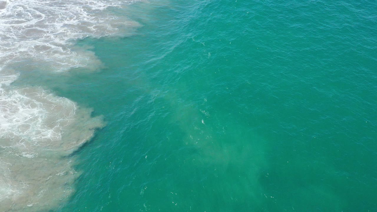 Top-down view of turquoise ocean in San Jose del Cabo with white foam breaking at the upper left and no visible people.