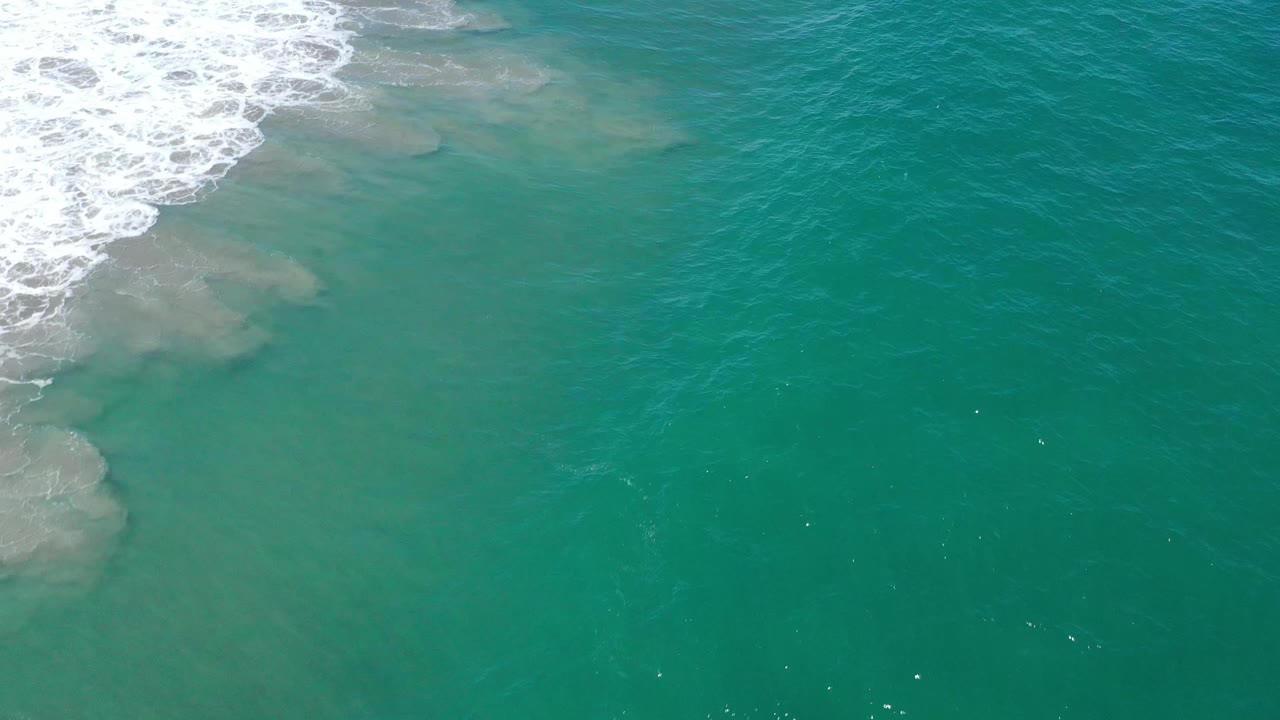 Turquoise sea near San Jose Del Cabo with white foam along the upper left and muted shallow patches beneath the water, no visible people.