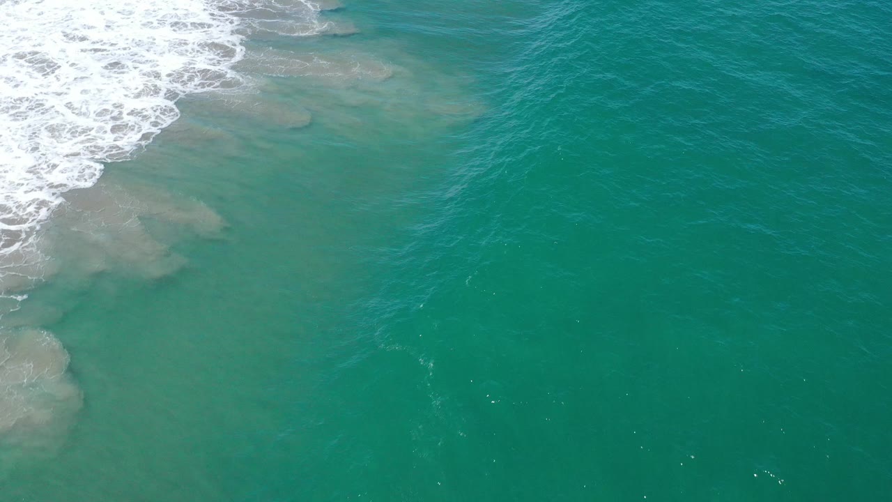 Open teal water in San Jose Del Cabo with faint underwater sand coloration and a small band of surf at the top left, no visible people.