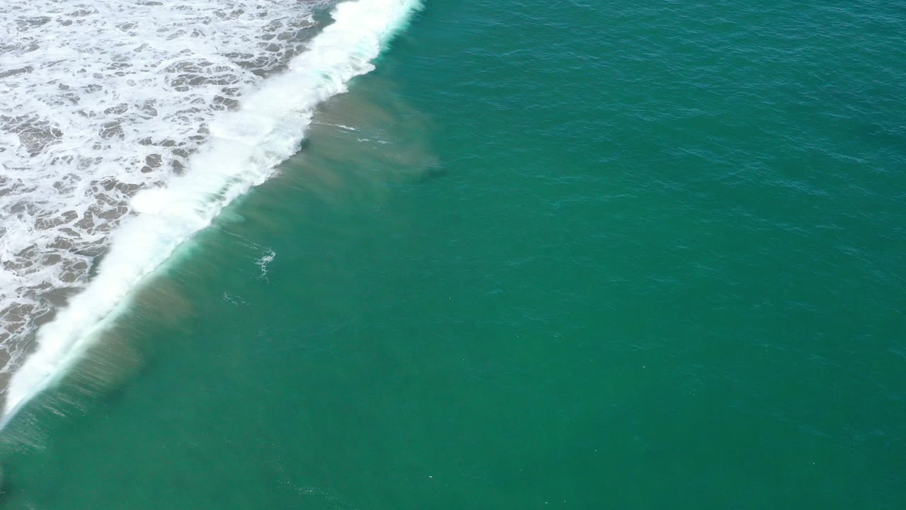 Overhead view of green water and a diagonal white surf line at San Jose Del Cabo, Mexico, with sandy color beneath the shallows and no visible people.