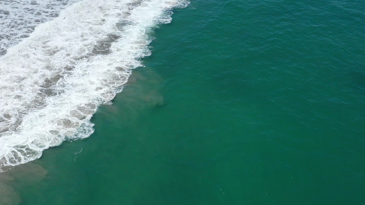 Overhead view of white surf rolling across teal water in San Jose del Cabo, Mexico, with pale underwater sand patches and no visible people.