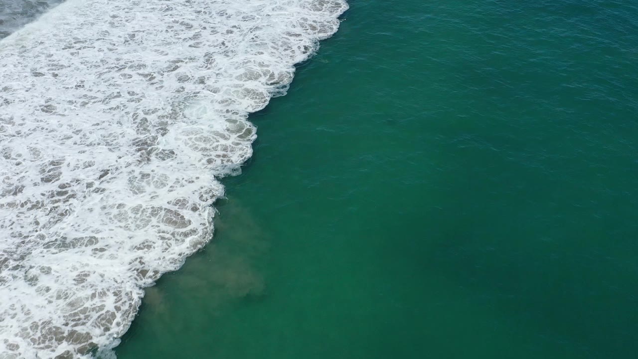 A foamy wave edge advances diagonally across green-blue water in San Jose del Cabo, Mexico, with cloudy sand visible below and no visible people.