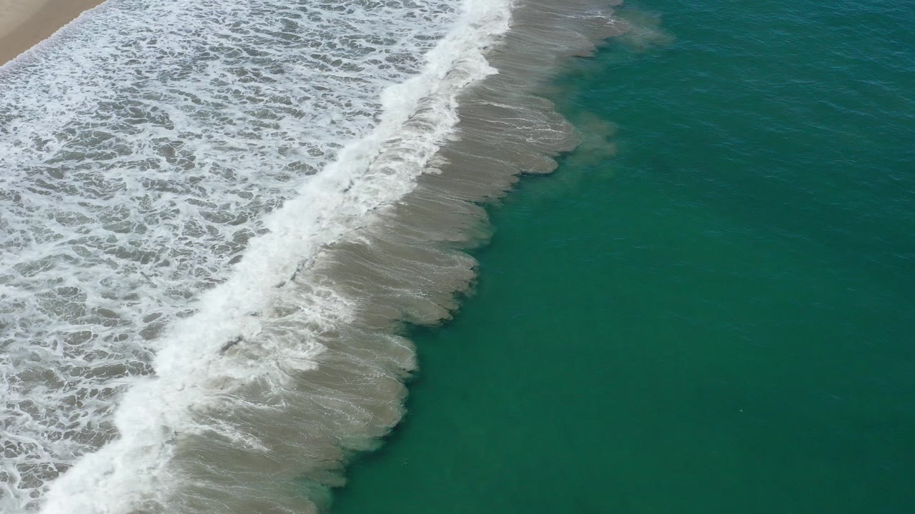 Overhead surf line breaking beside tan sand and green ocean.