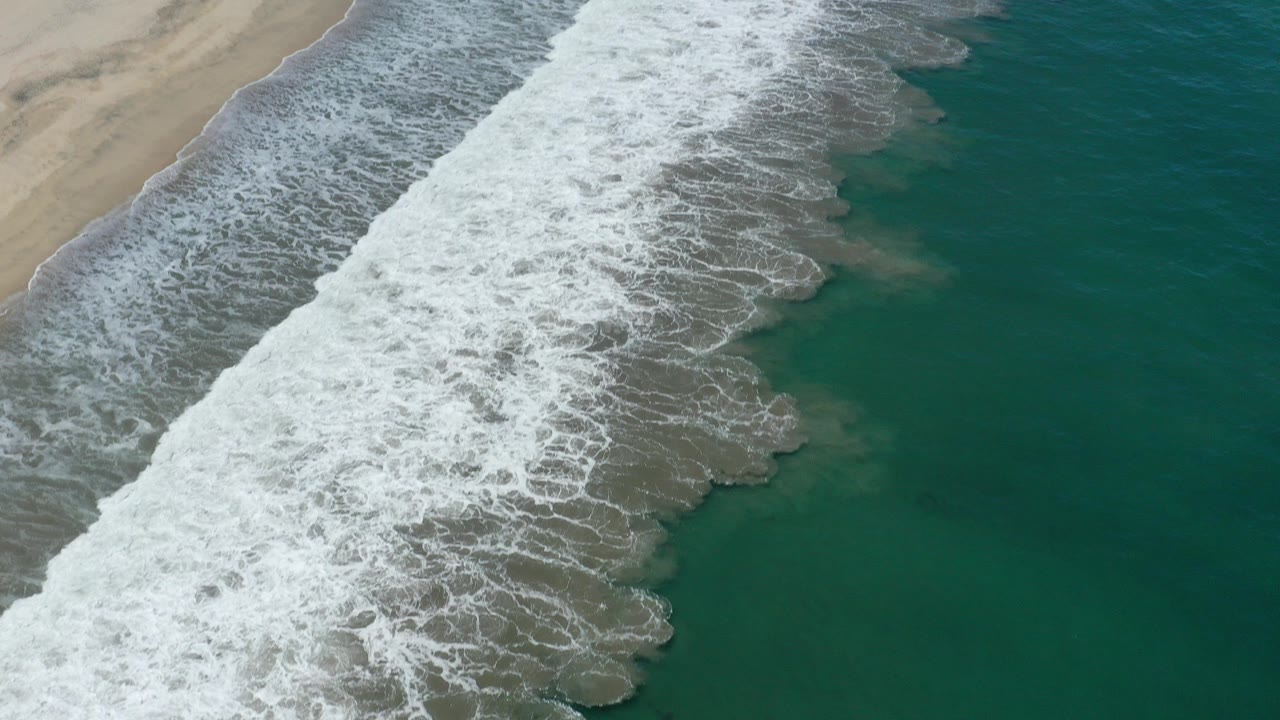 Aerial view of white waves breaking along a sandy beach beside green-blue water.