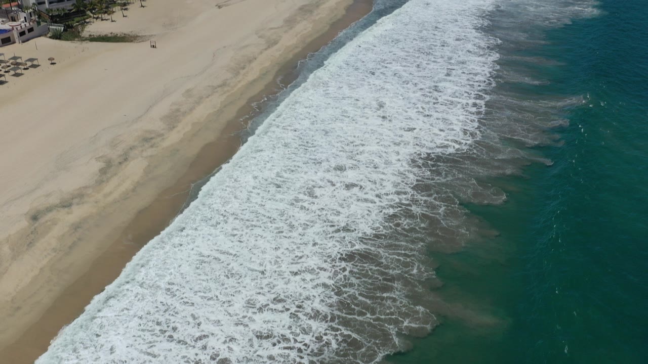 High aerial frame over San Jose del Cabo beach with pale sand at left, white surf crossing the center, turquoise ocean at right, resort edges near the top, and no visible people.