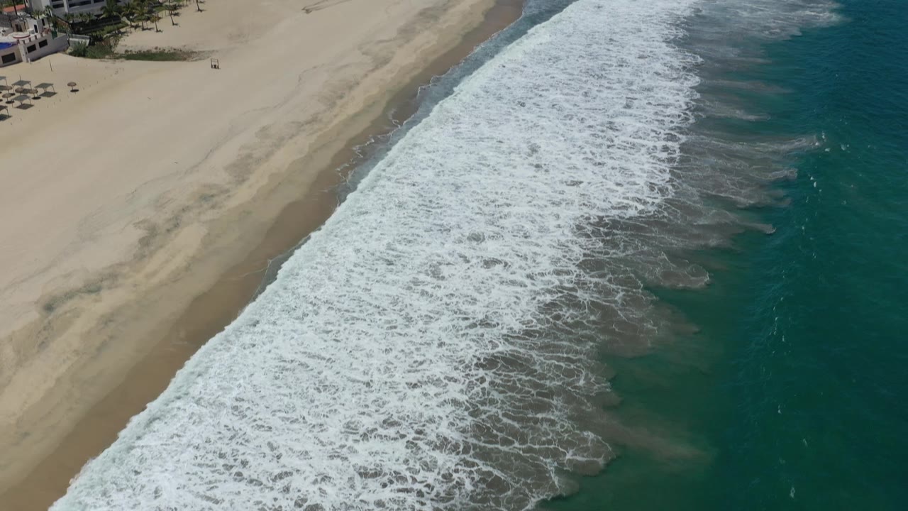 Aerial view in San Jose del Cabo with the breaking wave line spread across the middle, empty sand on the left, deep green-blue water on the right, and no visible people.