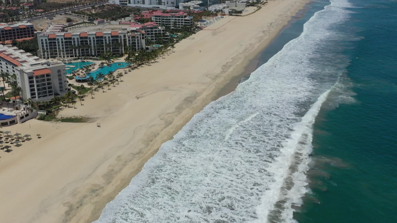Aerial view in San Jose Del Cabo showing resort buildings and pools at left, a wide sandy beach through center, blue-green ocean at right, and no visible people.