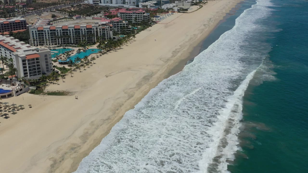 Aerial San Jose Del Cabo beachfront frame with hotel blocks and turquoise pools at left, pale sand in the middle, foamy surf bands at right, and no visible people.