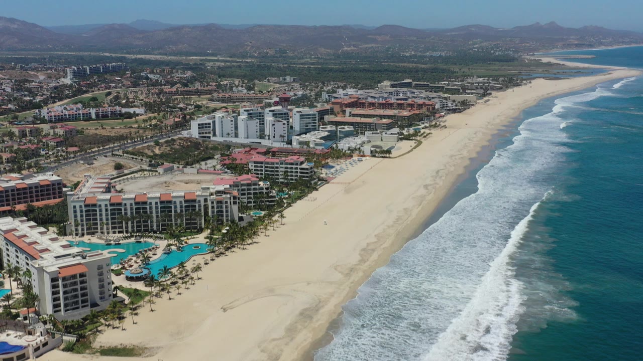 Aerial view over San Jose Del Cabo beachfront hotels and condos, with pale sand, blue surf, turquoise pools, inland roads, low hills, and tiny distant people on the beach.