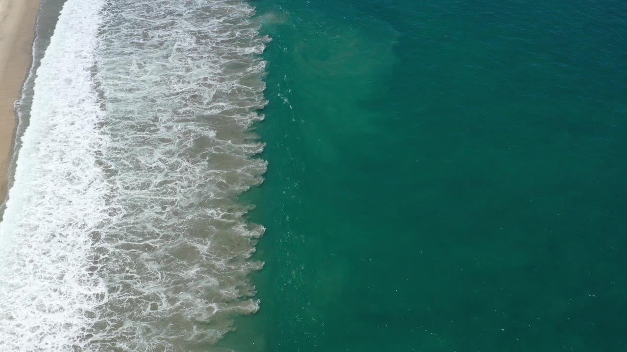 Overhead frame from San Jose del Cabo, Mexico showing white foam washing along tan sand at left and deep green ocean at right, with no visible people.