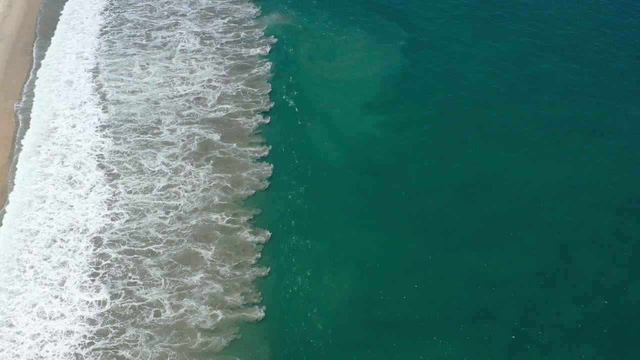 Overhead San Jose del Cabo, Mexico shoreline frame with dense white foam on the left and open green water across the right side, with no visible people.