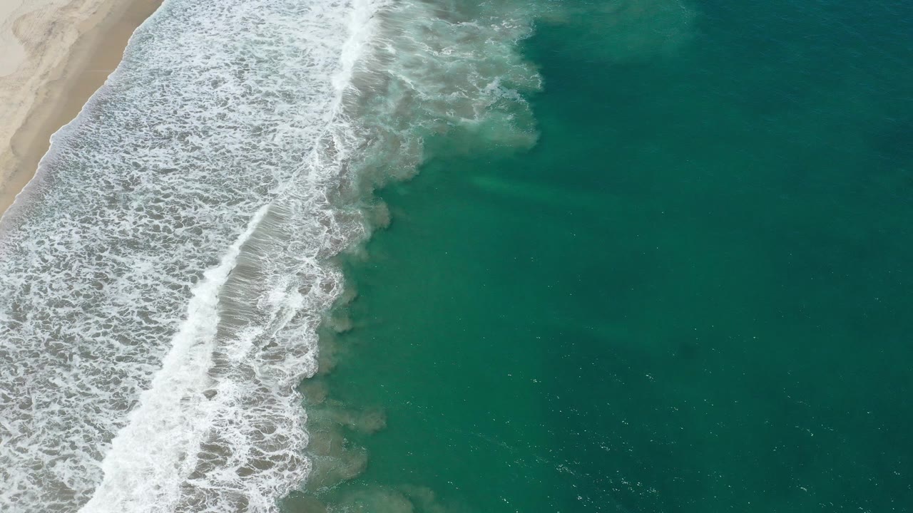 A later San Jose Del Cabo frame shows the white wave line shifted down the diagonal shoreline, with green water filling most of the right side and no visible people.