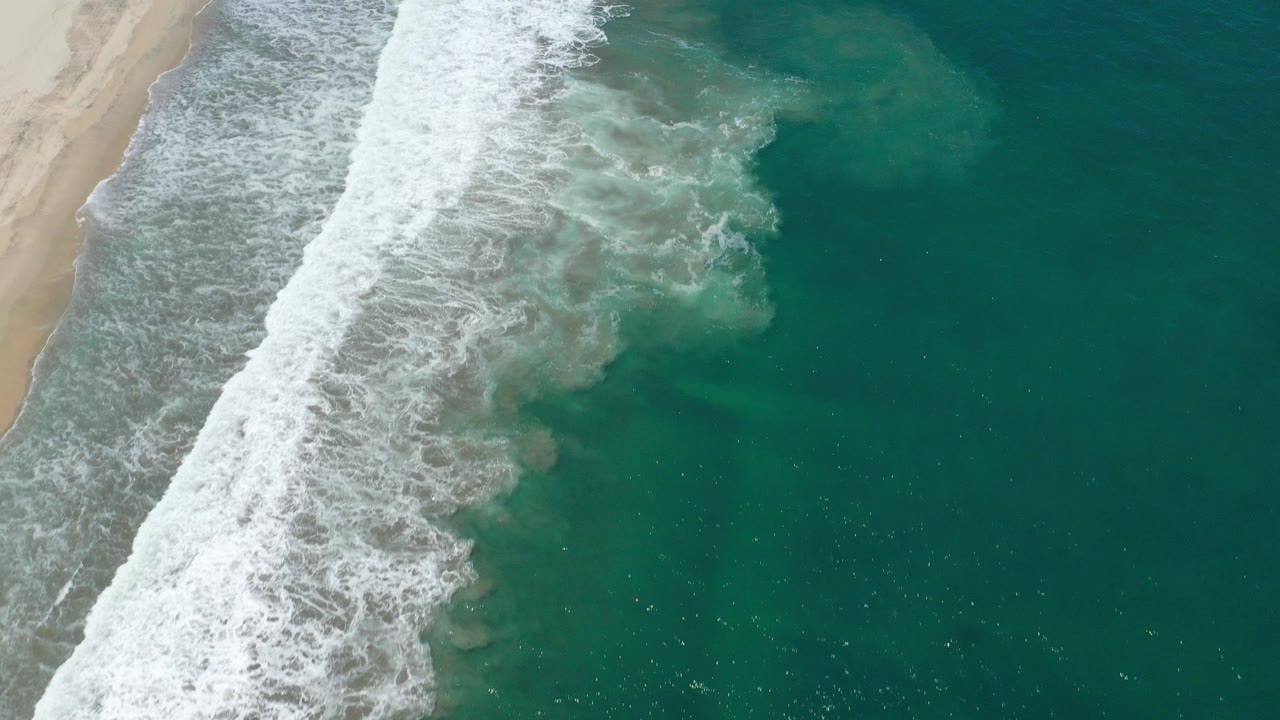 Overhead view in San Jose del Cabo with tan beach at left, a diagonal band of white surf, green-blue ocean to the right, and no visible people.
