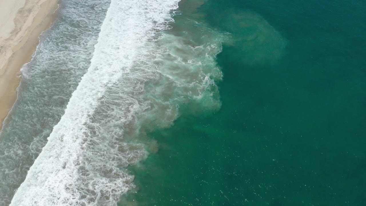 White surf curls beside open teal ocean from a vertical view.