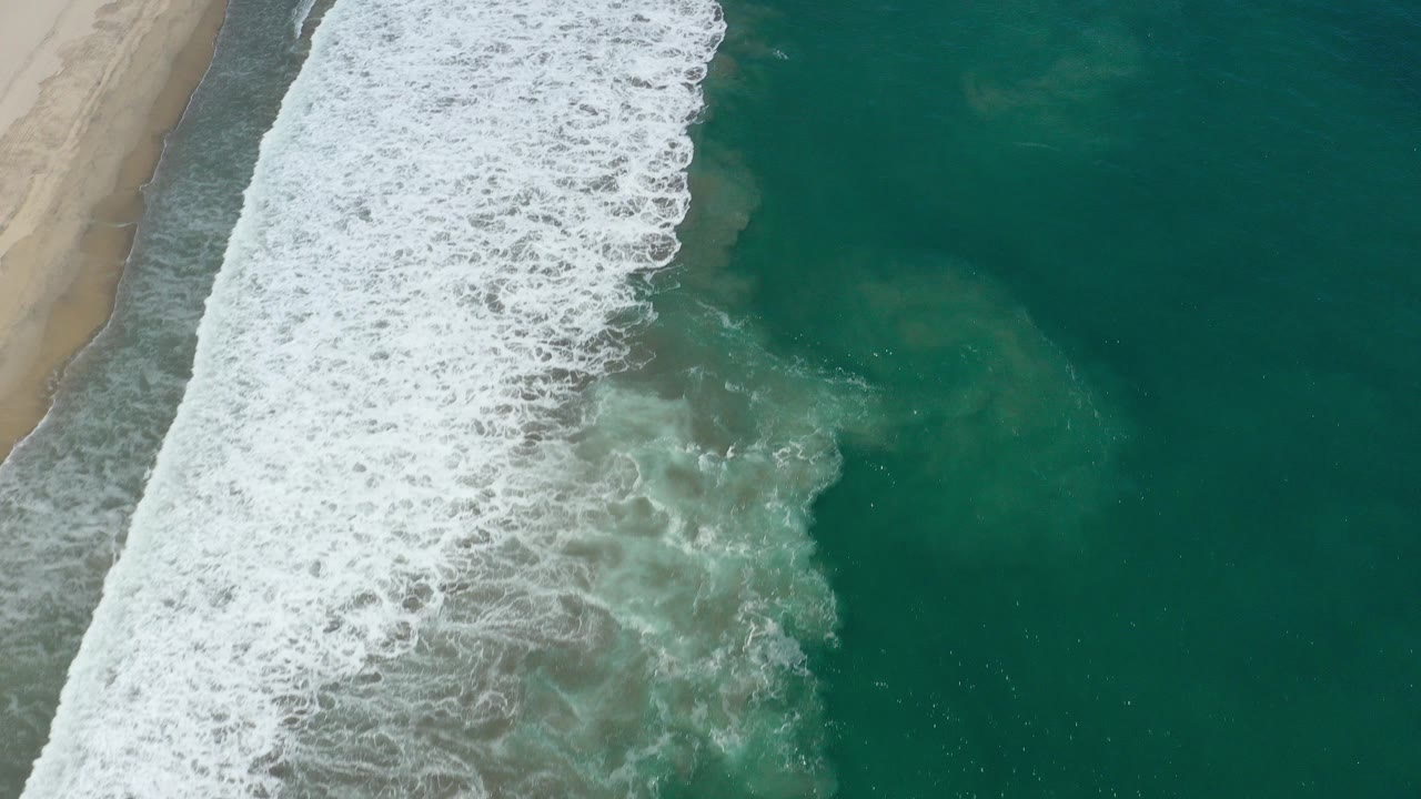 Overhead San Jose del Cabo shoreline view with white foam crossing diagonally beside green water and a narrow sandy beach at left, with no visible people.