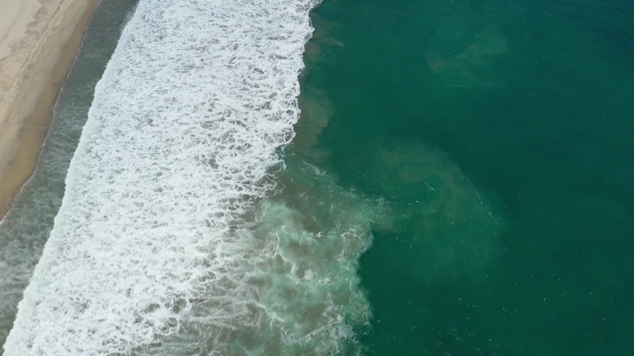 White surf breaks over green water with sand visible along the left edge.