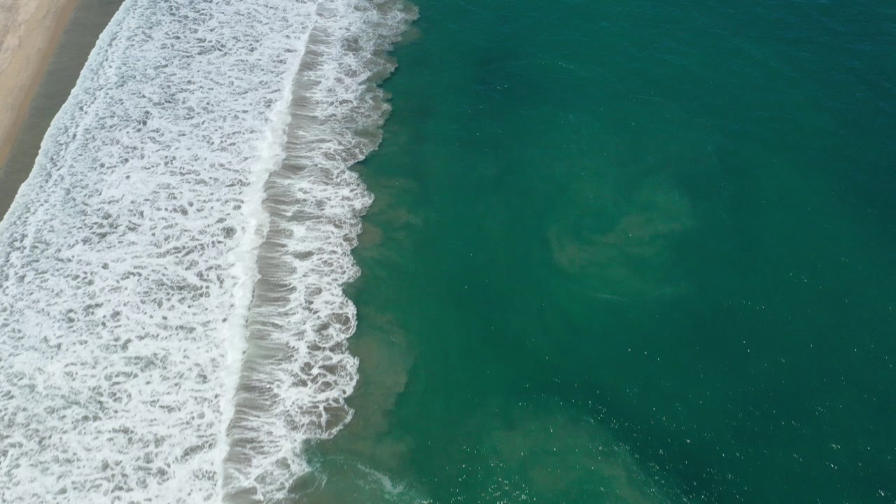 High overhead view of white seafoam breaking diagonally along tan sand and blue-green ocean in San Jose Del Cabo, Mexico, with no visible people.