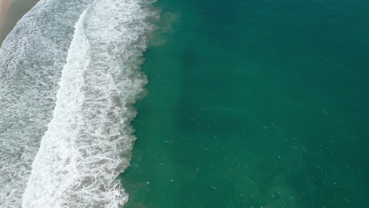 Overhead view in San Jose del Cabo, Mexico, where white surf enters from the left beside clear green-blue water, with no visible people.