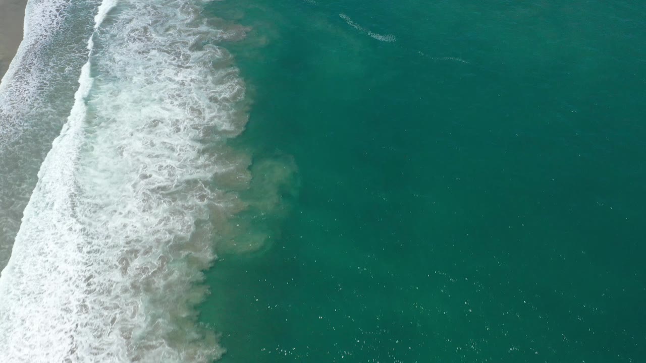 A breaking wave spreads white foam over green-blue water in San Jose Del Cabo, Mexico, while the right side remains mostly open water with no visible people.