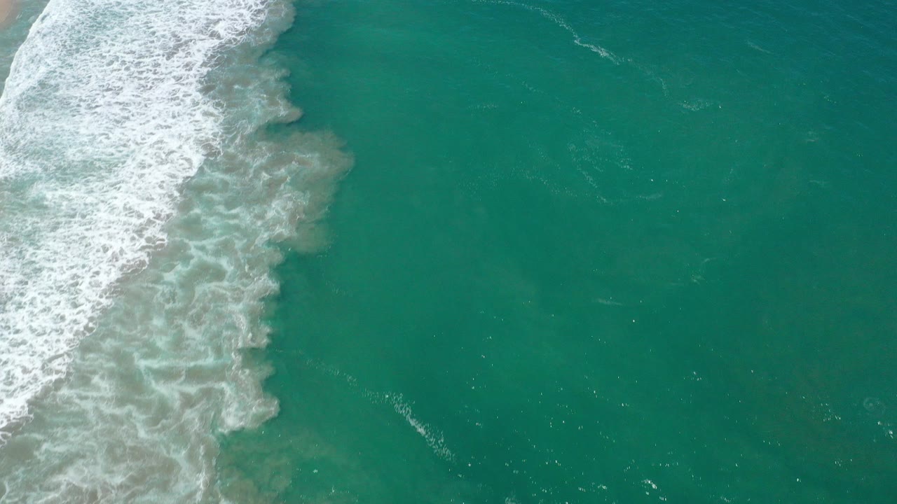 Overhead view of San Jose del Cabo ocean water with white surf entering from the left, turquoise water across the frame, and no visible people.