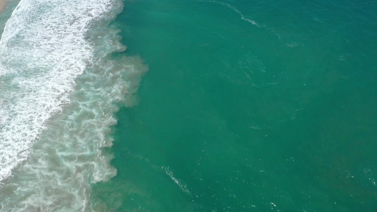 San Jose del Cabo shoreline water seen from above, with foamy surf concentrated on the left and broad green-blue ocean to the right with no visible people.