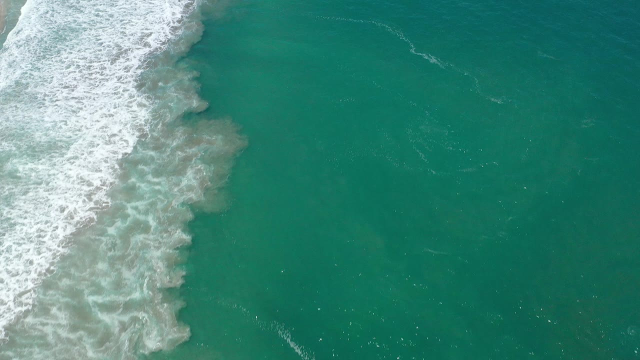 Aerial ocean frame with surf at upper left and clear water filling the right side.