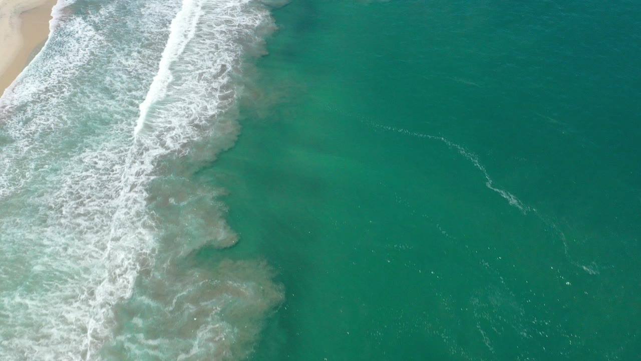 Overhead view of white surf breaking beside tan sand and turquoise water in San Jose del Cabo, Mexico, with no visible people.