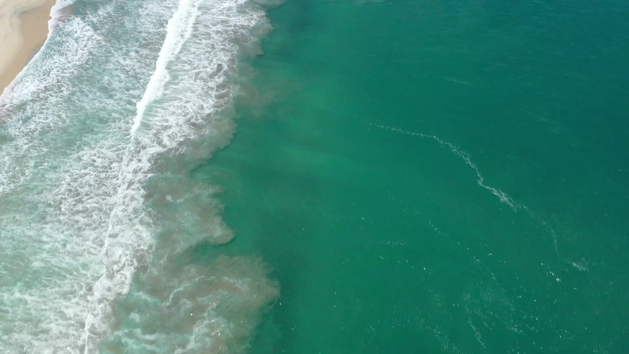 White foam spreads farther down the left shoreline while green-blue ocean fills the right side in San Jose del Cabo, Mexico, with no visible people.