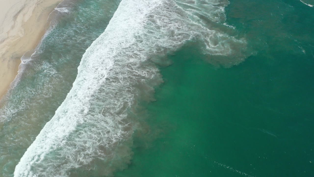 Aerial view of teal ocean water and white surf breaking diagonally beside sandy beach in San Jose del Cabo, Mexico, with no visible people.