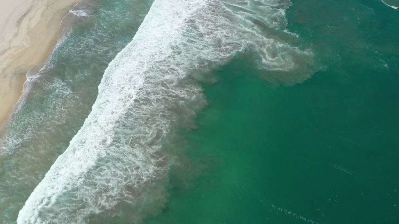 White foam spreads farther across the teal shoreline water near the sandy edge of San Jose del Cabo, Mexico, with no visible people.