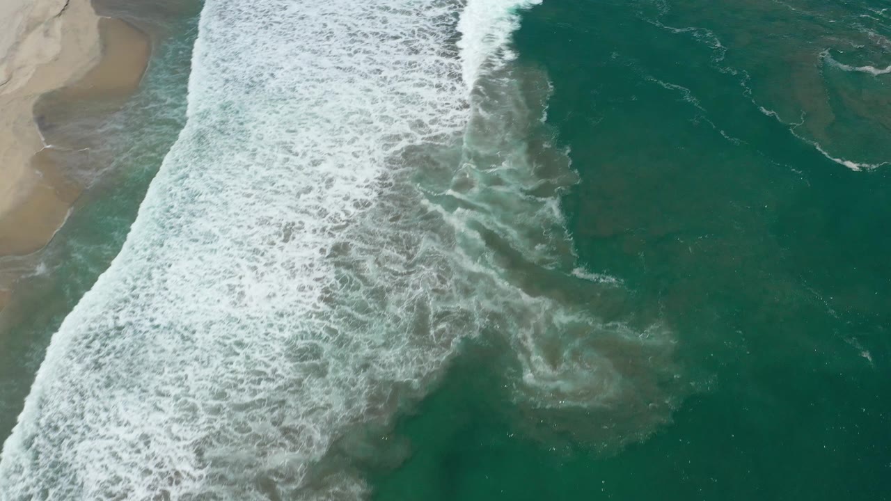 Aerial San Jose del Cabo frame with a broad white surf band spreading diagonally across teal water, sand partly visible at the upper left, and no visible people.