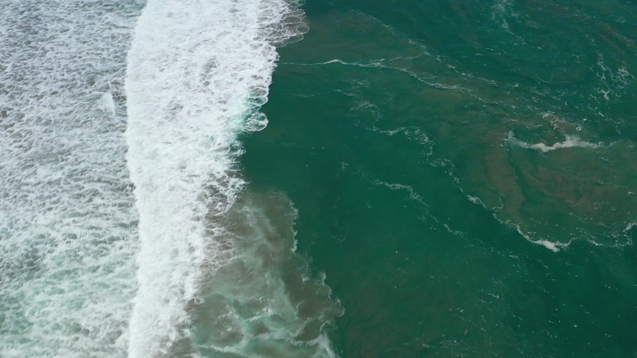 Overhead view of teal ocean in San Jose Del Cabo, Mexico, with a broad white surf line entering from the left and no visible people.