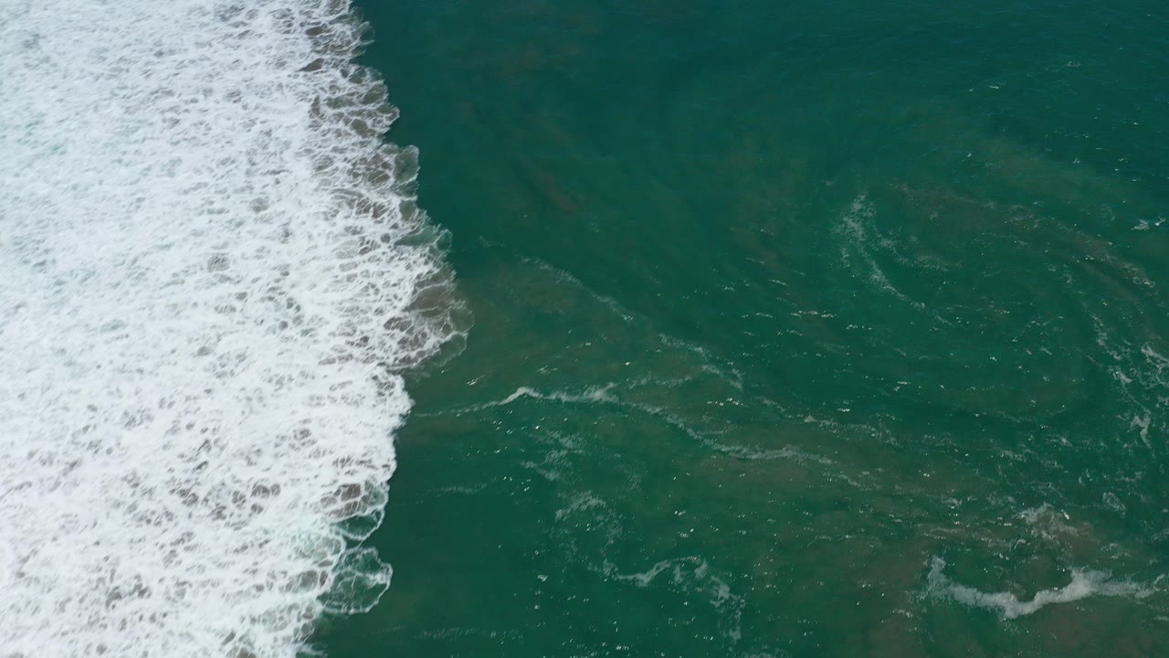 Aerial view over San Jose del Cabo ocean water, with a broad strip of white surf along the left side and no visible people.