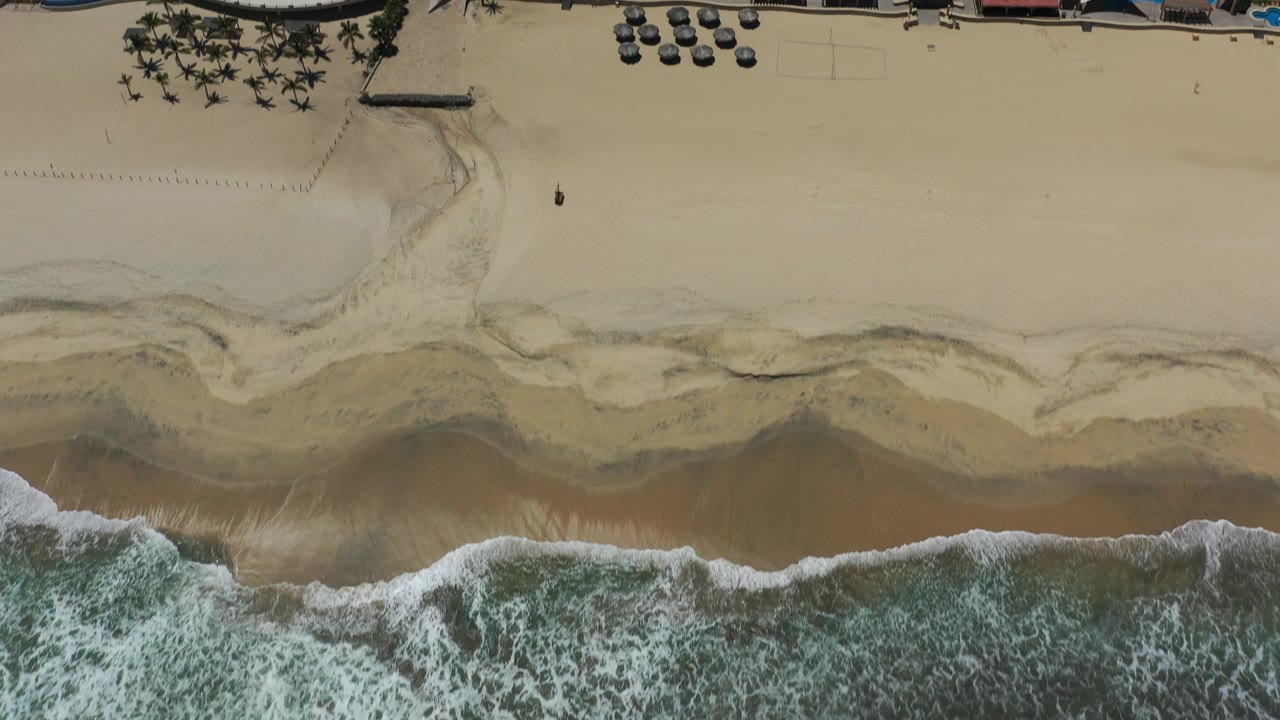 Overhead view of surf reaching a wide sandy beach beside resort grounds.