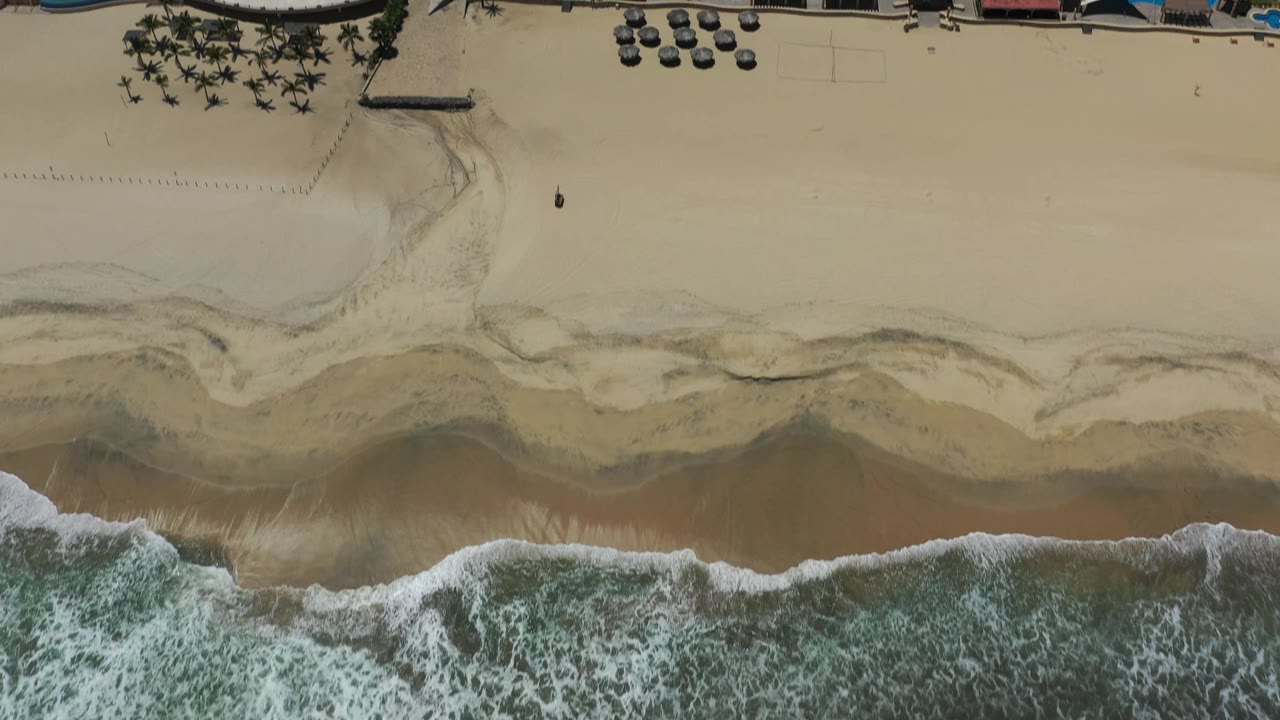 Overhead San Jose Del Cabo beach frame showing a scalloped line of surf below pale sand, dark water marks, beach umbrellas near the resort, palm rows, pools, and small distant people on the beach.
