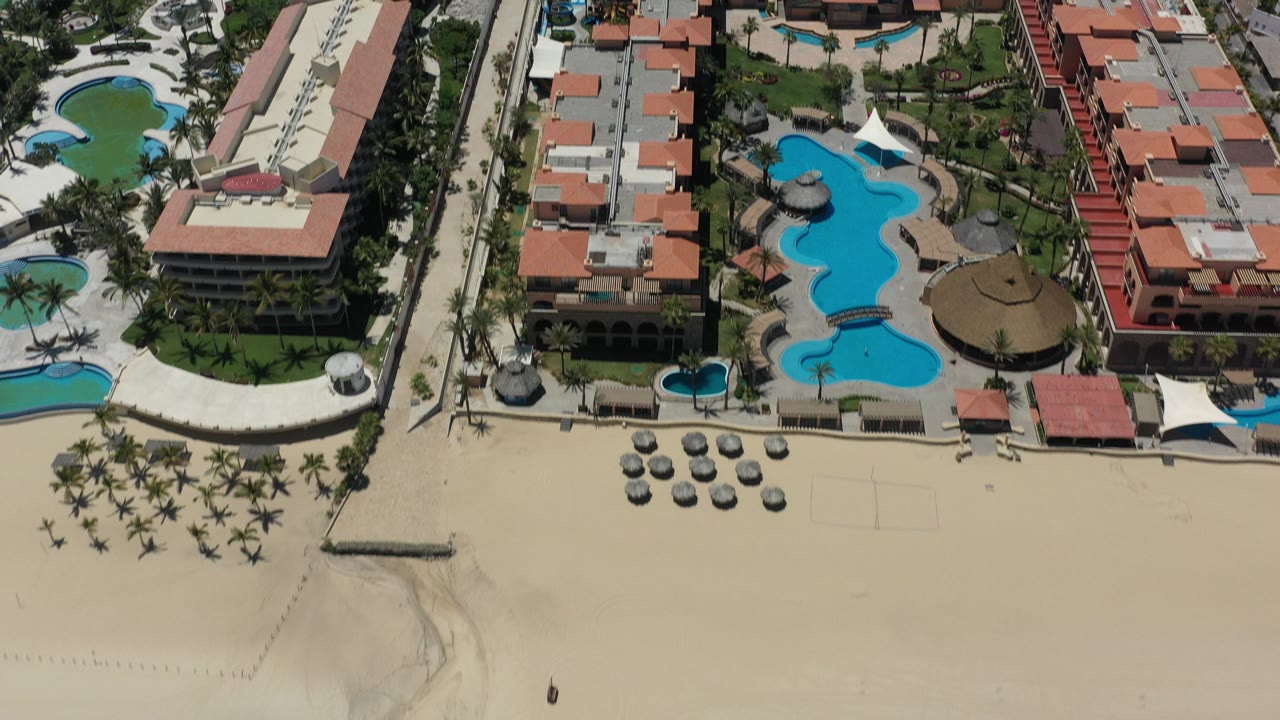 Aerial view in San Jose Del Cabo showing red-roofed resort buildings, turquoise pools, palm trees, umbrellas on beach sand, and no visible people.