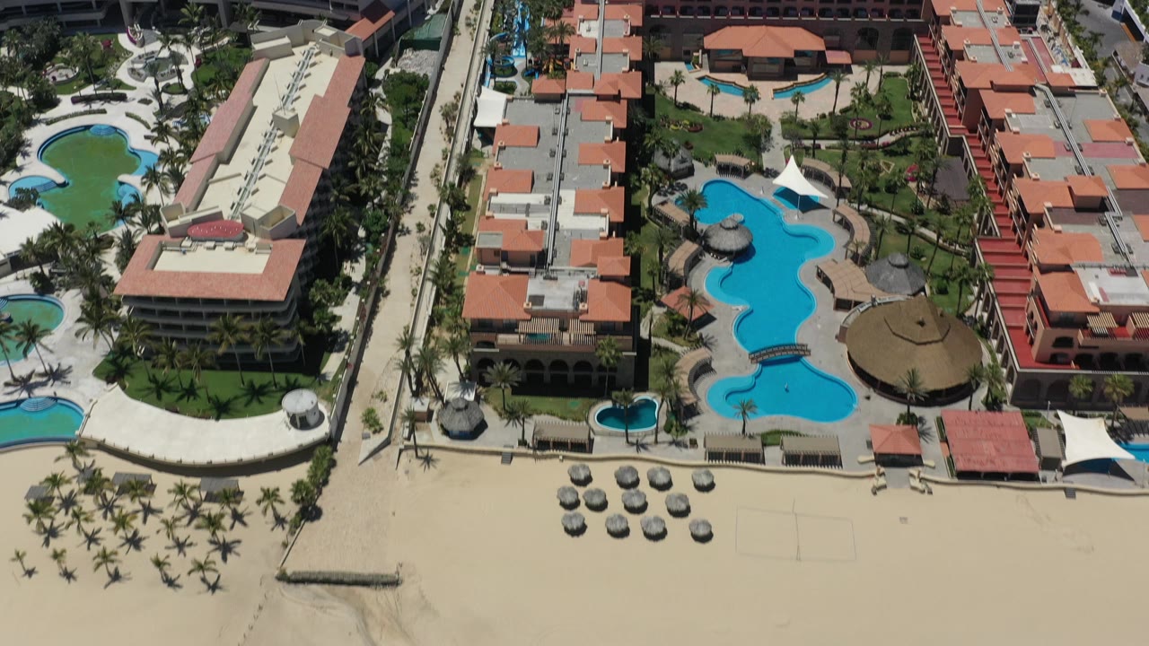 Drone-style view of pools, palms, rooftops, and empty beach area.