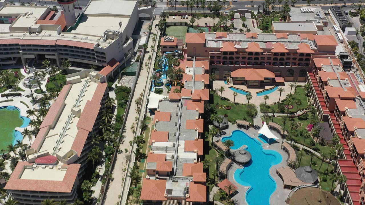 Aerial view over a San Jose del Cabo resort complex with terracotta roofs, blue swimming pools, palm landscaping, adjacent roads, and no visible people.