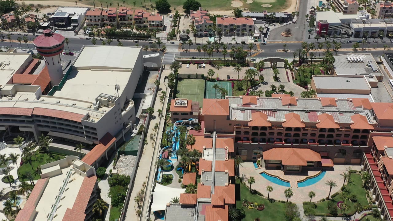 Aerial top-side view in San Jose del Cabo with the pool courtyard centered below red-tile roofs, tennis courts at left, roads beyond, and no visible people.