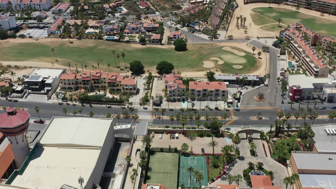 Second aerial frame in San Jose del Cabo with the road and tennis courts lower in frame, red-roofed buildings near center, and the grass field stretching behind them with no visible people.