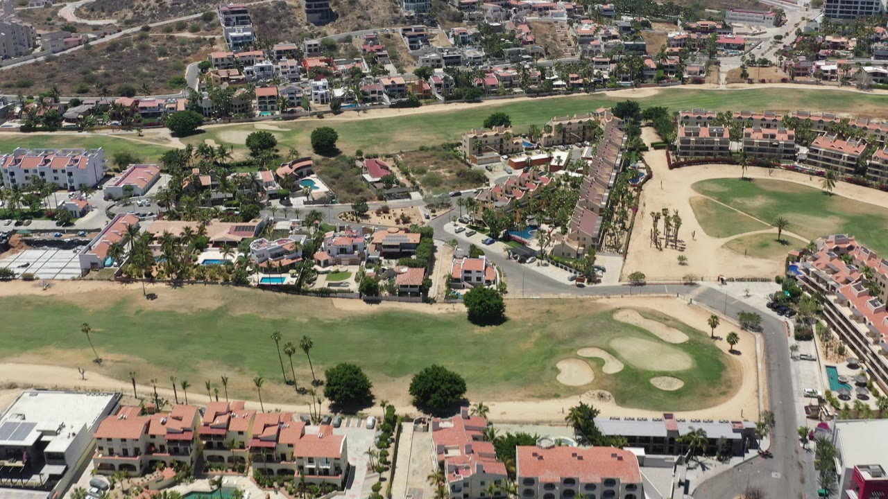 High aerial view over San Jose del Cabo showing green golf fairways, sand traps, terracotta-roof homes, roads, dry hills, and no visible people.