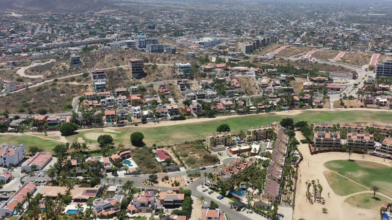 Aerial frame in San Jose del Cabo with residential blocks across dry terrain, a curved road near the hillside, green field sections at the bottom edge, distant mountains, and no visible people.