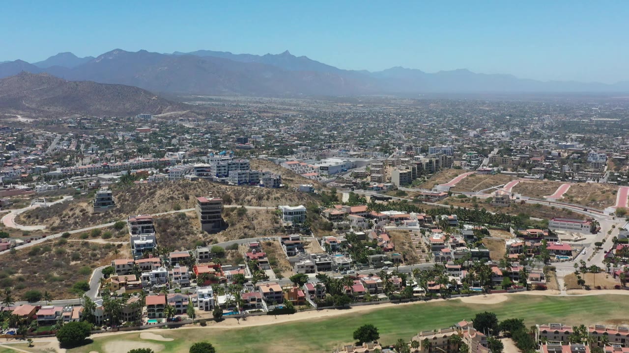 Wide San Jose del Cabo aerial frame showing residential blocks, winding roads, dry hillside lots, mid-rise buildings, hazy mountains, blue sky, and no visible people.